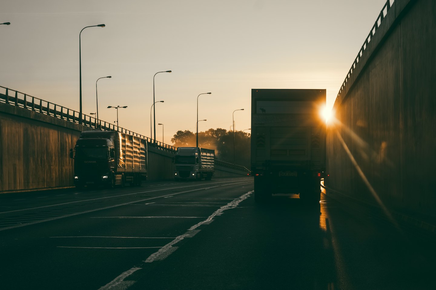 Semi-trucks drive along a multi-lane highway at sunset.