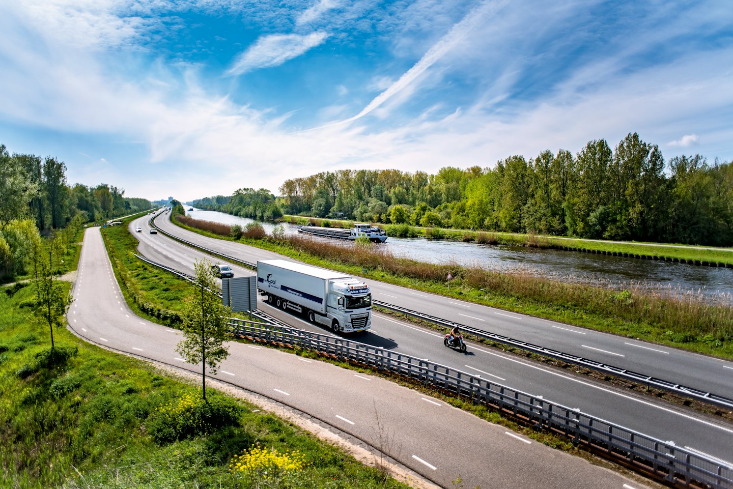 A semi-truck and motorcycle travel along a highway beside a canal.