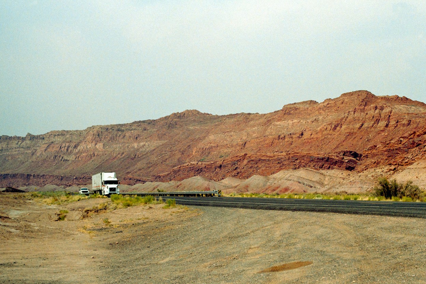 A semi-truck drives on a desert highway near red rock formations.